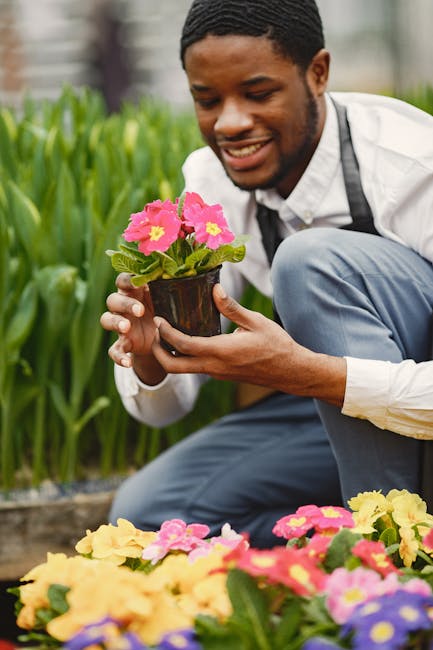 A gardener wearing a grey long-sleeve shirt with reflective stripes, dark trousers, and a wide-brimmed hat is walking along a curved, paved pathway inside a greenhouse. They are holding a small garden
